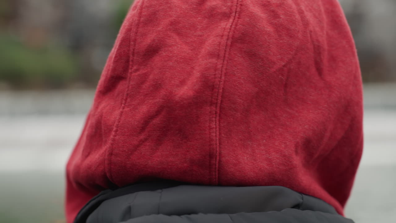 Persona con sudadera roja junto al río mirando hacia arriba, gafas, gorra amarilla, pelo rizado, chaqueta acolchada negra, puente de madera de fondo, cascada suave, luz nublada, bokeh cinematográfico, estado de ánimo reflexivo.