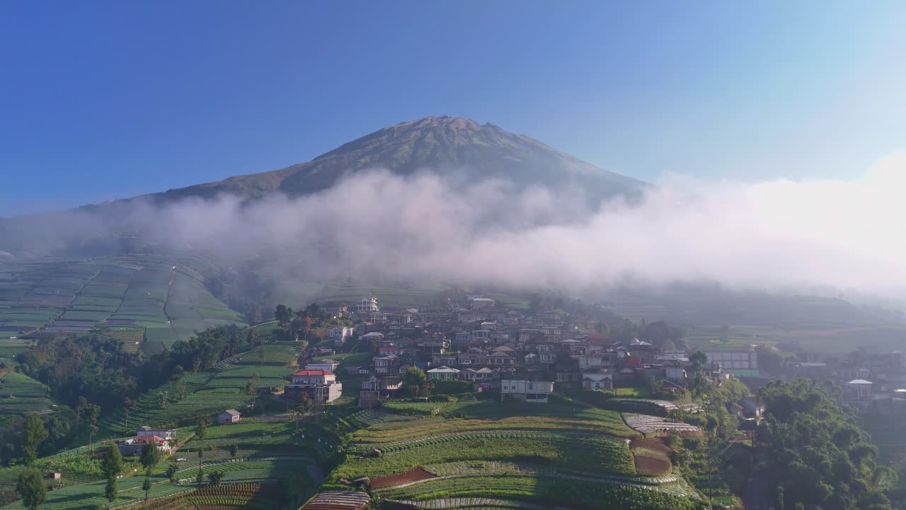 Vista aerea con drone di un pendio montuoso con campi terrazzati, case di paese e un'imponente montagna coperta dalle nuvole