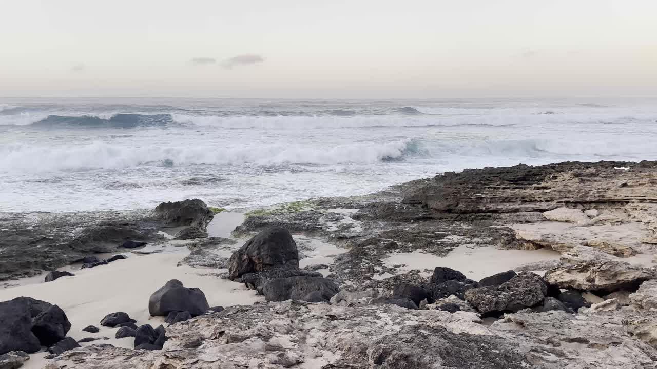 The powerful waves of the Pacific Ocean crash against the rugged rocky shoreline at dusk in Oahu, Hawaii