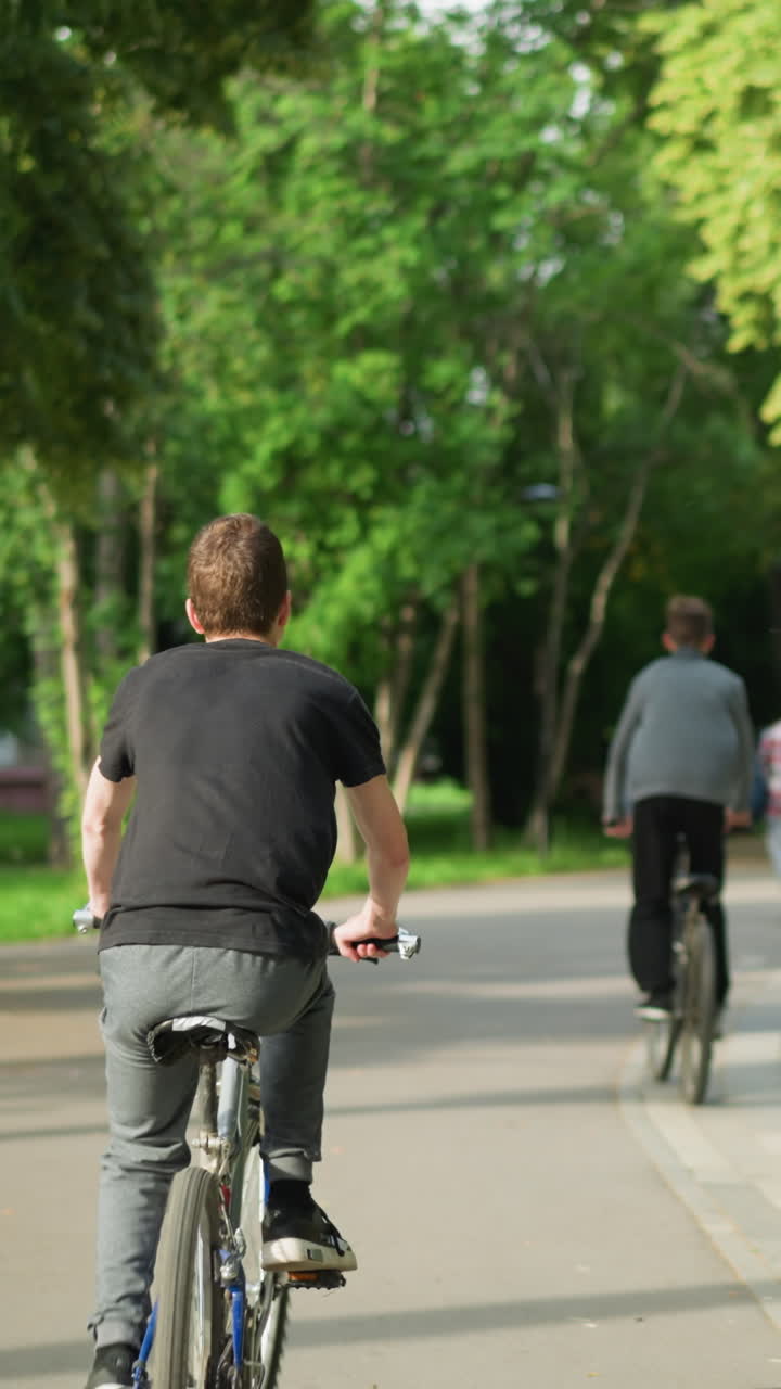 두 형제는 개방된 공간에서 자전거를 타고 다니며, 누군가의 흐릿한 시야가 그들 앞에서 걸어가는 것을 볼 수 있습니다. 배경에는 스쿠터를 타고 있는 어린 소년도 약간 흐릿하게 보입니다.