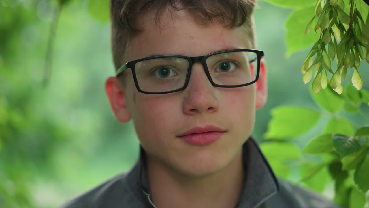 Close-up of young boy with pink lips, wearing glasses and a grey top, looking directly at the camera with a slight smile, he is surrounded by blurred greenery