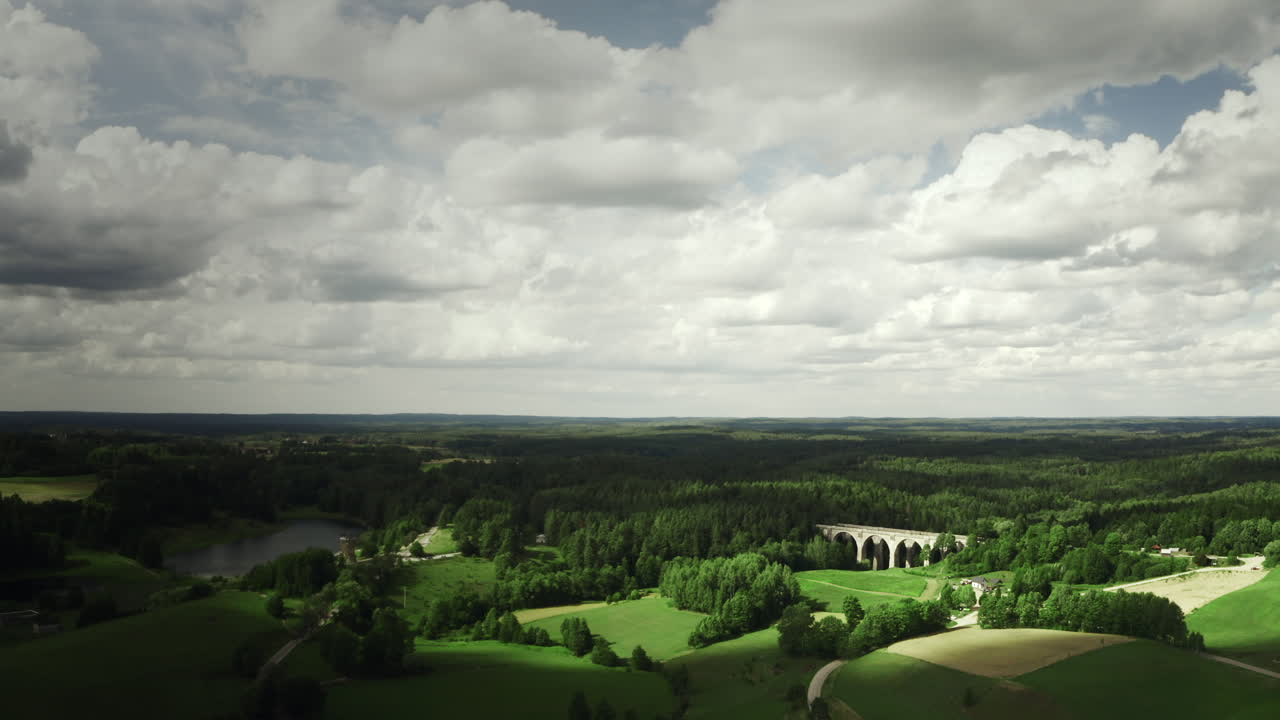 Drone shot of historic bridges. Lush green landscape with rolling hills, forests, lake and old stone viaduct under cloudy sky. Stanczyki Bridges during summer. Brilliant lights, moving shadows.