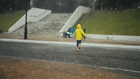 happy teenage girl in a yellow jacket and orange rubber boots runs through large puddles during heavy rain in the park