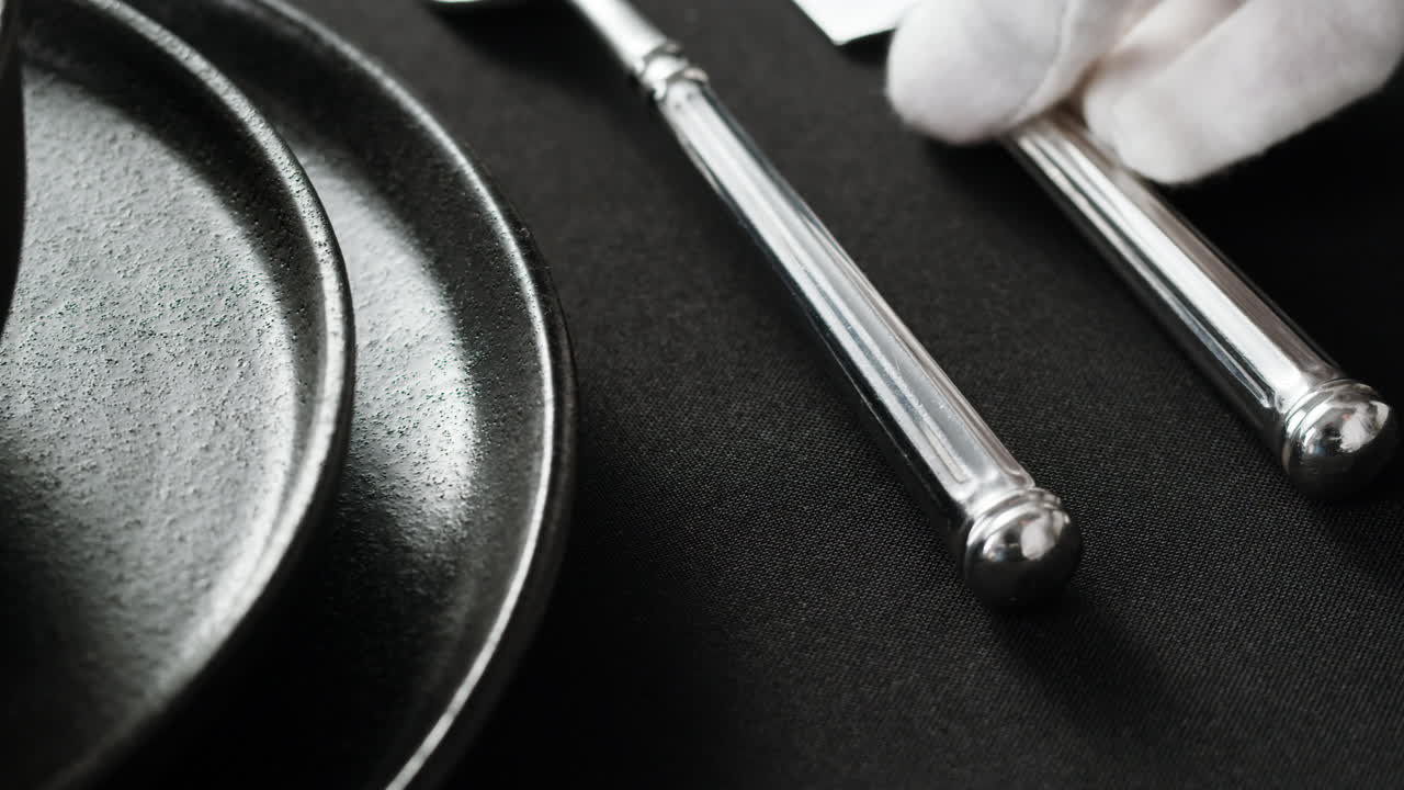 Minimalist table setting with white plate, fork, and knife arranged neatly, and a gloved hand adjusting cutlery.