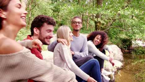 Five-young-adult-friends-taking-a-break-during-a-hike-sit-talking-by-a-stream-in-a-forest,-handheld