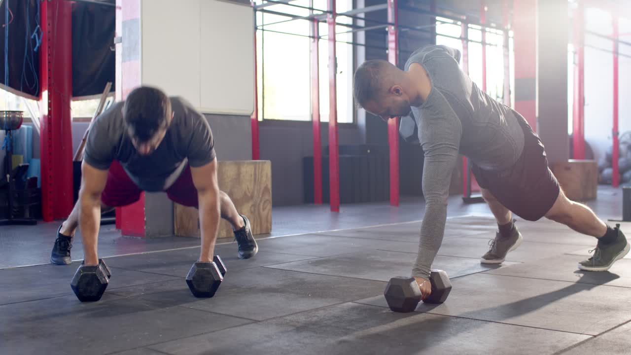 Dúo masculino diverso entrenando en el gimnasio, haciendo planchas que pasan a flexiones y remo con mancuernas para fortalecer