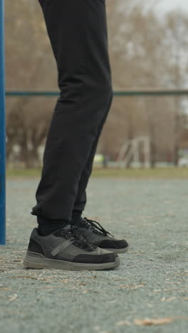 close-up of a person's leg in motion, swinging near a blue pole in a stadium, in the background, a boy plays football on a field with cars parked in the distance