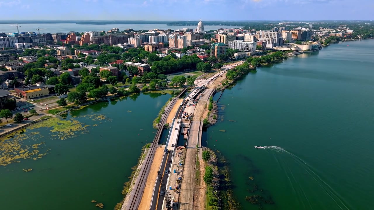 Hyperlapse aereo con drone in volo sopra una strada rialzata sul lago e una ferrovia in costruzione, che conduce verso lo skyline del centro città e l'edificio del Campidoglio dello Stato a Madison, Wisconsin, in una giornata di sole