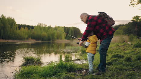 curious little boy is learning to fish on coast of forest lake grandfather is helping to grandson