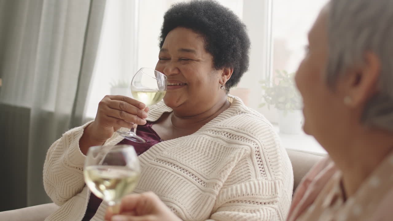 Tilting up of joyful mature African woman sitting on sofa at home, drinking white wine, talking and laughing with unrecognizable friend