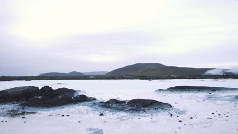 Close-up-of-rocks-in-a-frozen-lake,-cars-driving-on-the-shore,-Iceland