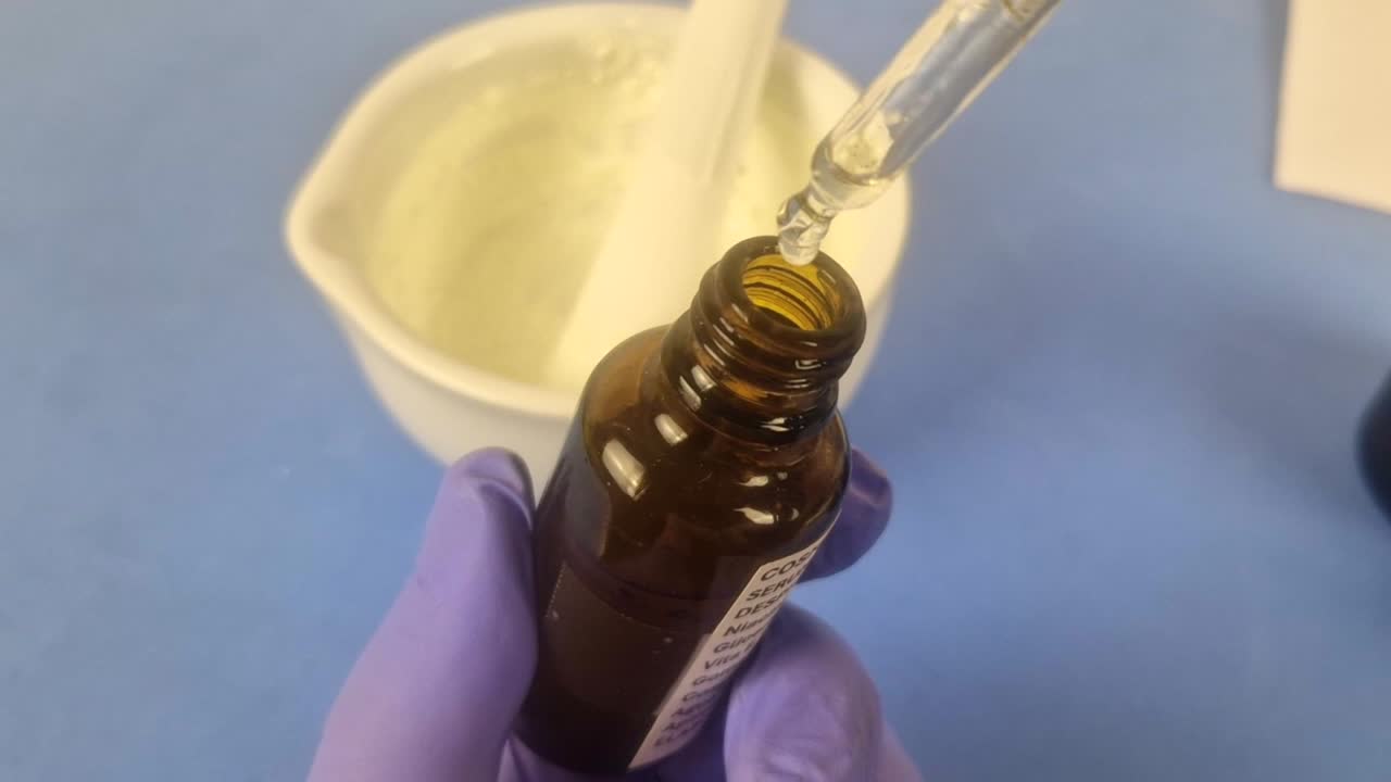 Macro shot of a clear cosmetic serum dripping from a glass dropper into an amber bottle. Shows pharmacist's gloved hands in a clean laboratory, highlighting precision