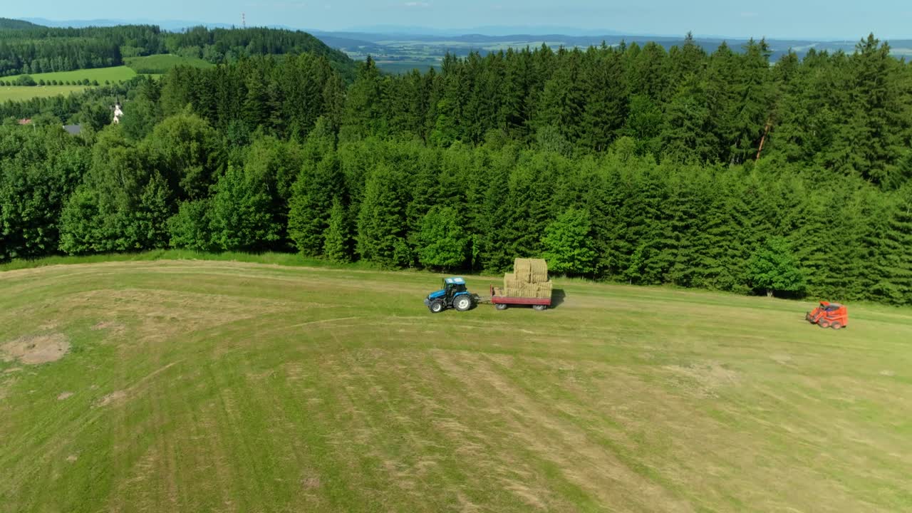 Aerial drone view of a tractor harvesting golden hay bales across vast fields, showcasing summer harvest in a picturesque rural landscape.