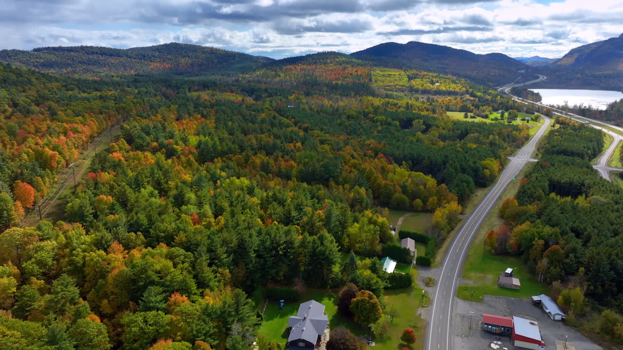 Few cottages near the highway crossing the picturesque autumn forest. Mountains under the cloudy sky at backdrop.