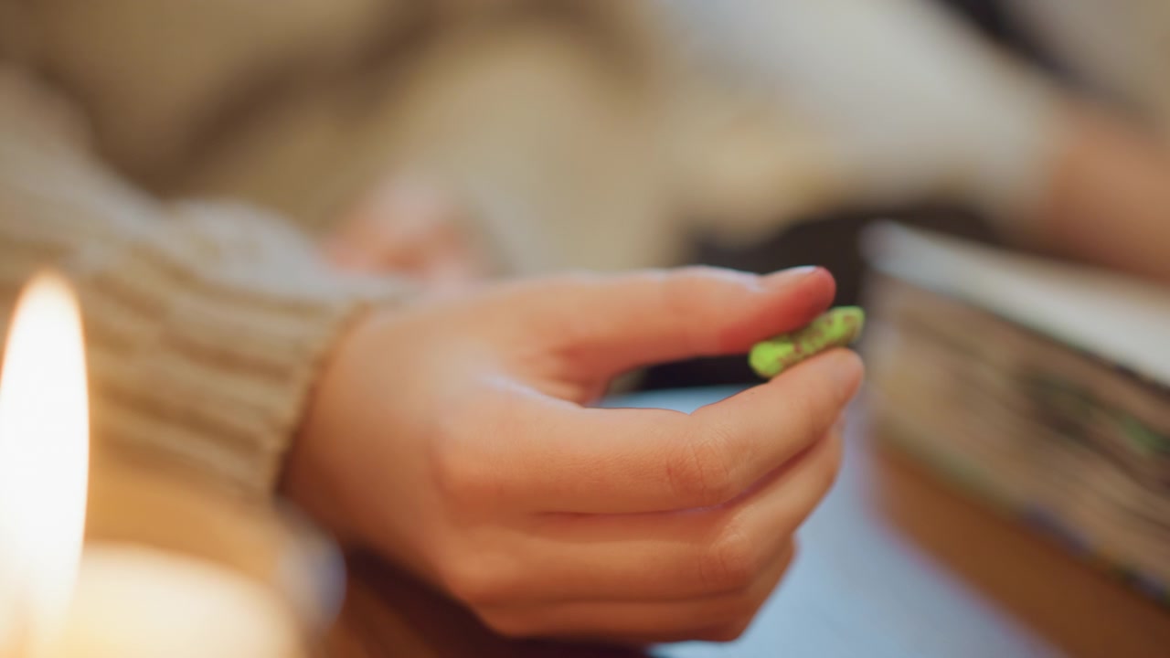 Primo piano di una mano che tiene un pastello verde sopra un tavolo con libri e una candela. La mano rimane ferma mentre lo sfondo mostra una messa a fuoco morbida sui materiali di studio. L'illuminazione calda proveniente dagli oggetti vicini crea.