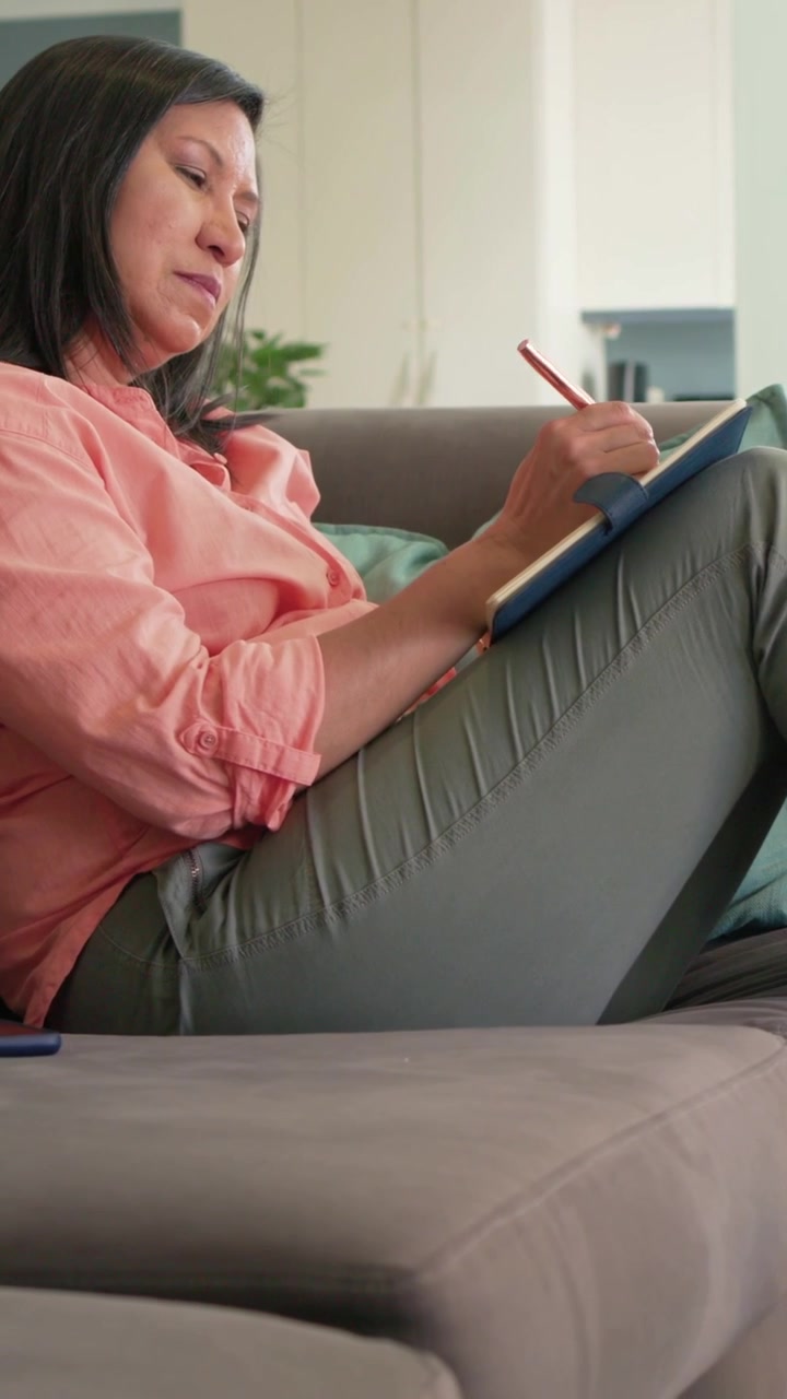 Video verticale: Donna in camicetta color corallo con una matita alla bocca che scrive un diario in un quaderno blu a casa.