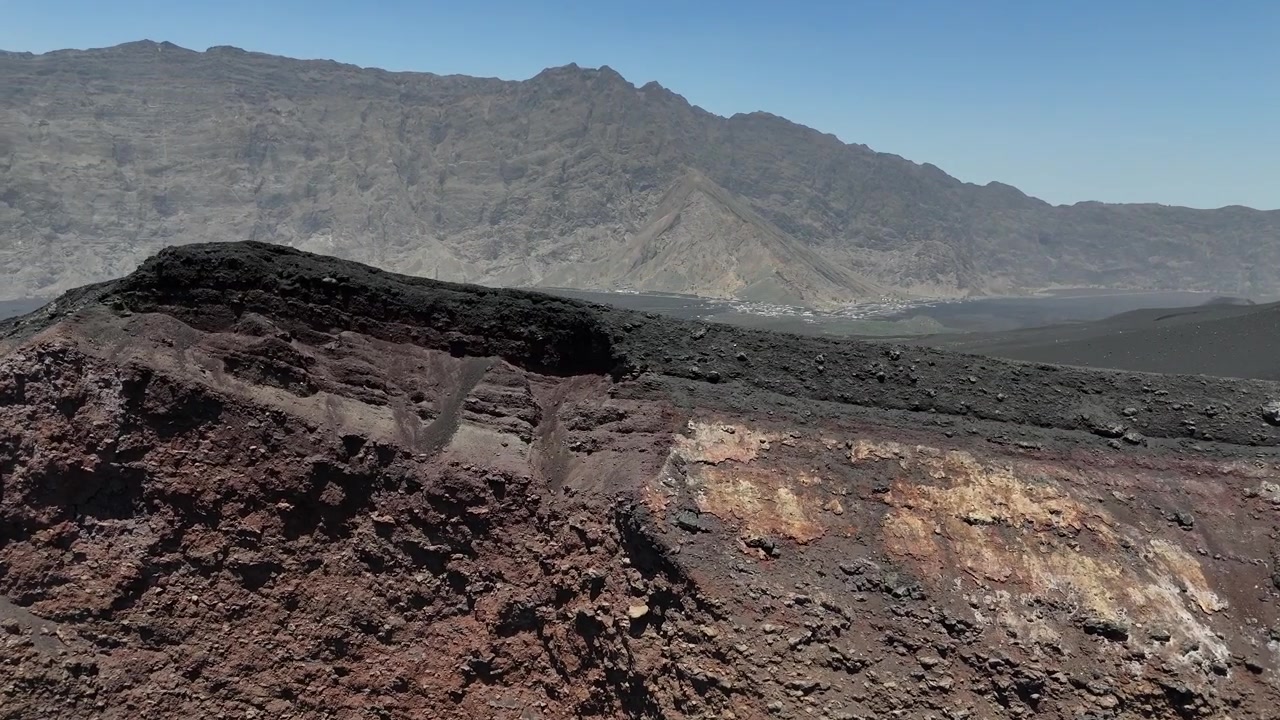 Ripresa ravvicinata con drone del bordo del cratere vulcanico di Pico do Fogo, che mostra le rocce basaltiche scure e porose e la vasta pianura di Chã das Caldeiras con la parete di Bordeira in lontananza