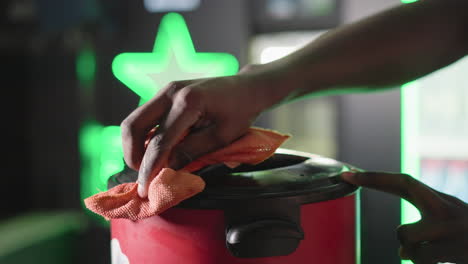 bartender wiping red trash bin with orange cloth behind bar counter. green neon heineken star sign illuminates the background, adding to the festive atmosphere. close-up of hand and bin