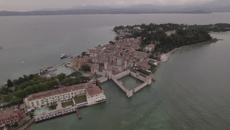 Toma-De-Drone-De-Simione-Italia-Mirando-La-Antigua-Fortaleza-En-Un-Día-Gris-Con-Barcos-En-El-Agua-Cerca-Del-Lago