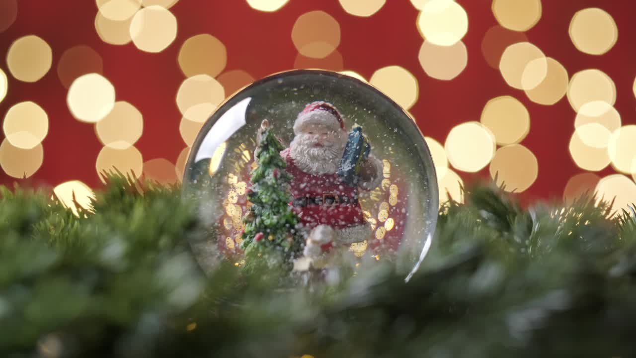 globo de nieve de navidad con santa claus y la nieve con ramas de abeto girando lentamente en cámara lenta contra un fondo rojo con bokeh parpadeando luces de navidad en la noche de navidad.