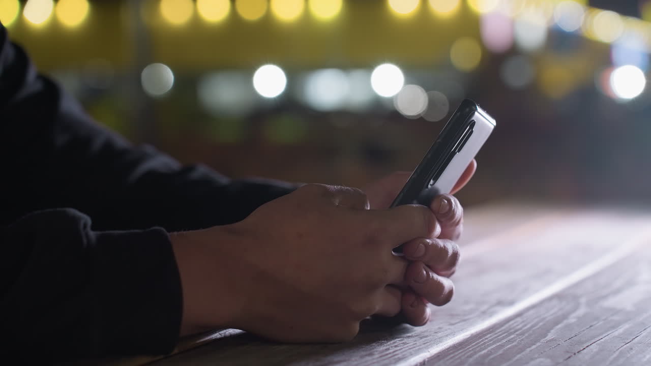 Hand view of person holding phone on wooden table while another hand reaches in from behind causing phone to be dropped under soft night lighting with warm bokeh city background