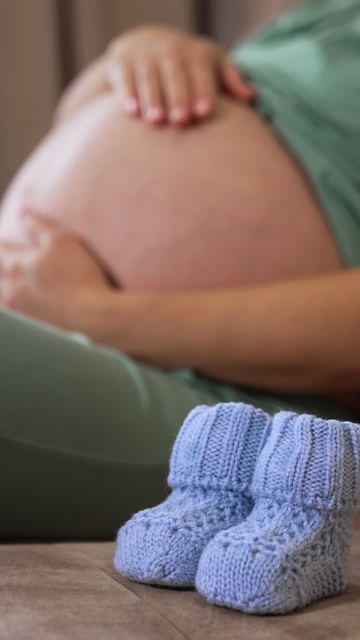 Pregnant woman with big bare belly sitting on the couch. Lady caressing her pregnant belly tenderly and then putting baby socks on it. Vertical video