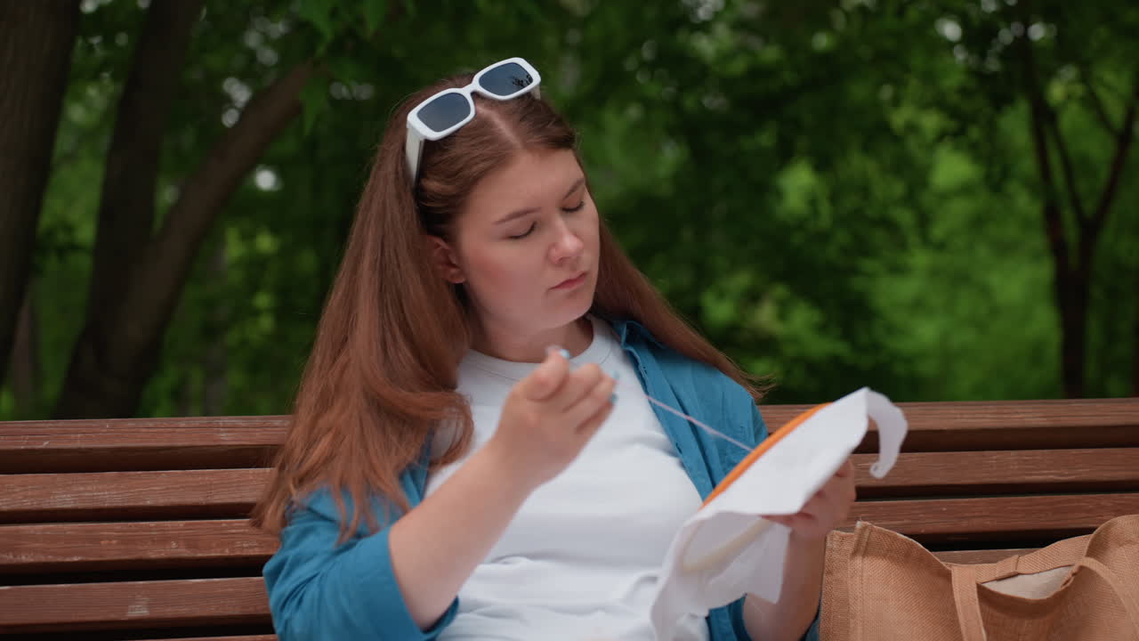 Young designer sitting on wooden park bench concentrating on embroidery work, holding needle and fabric hoop, wearing casual blue shirt and white outfit