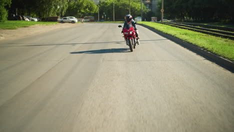 dos parientes viajan en una bicicleta eléctrica con cascos, navegando por una carretera, se ve un coche de color ceniza delante de ellos mientras viajan, con árboles y postes eléctricos en el fondo