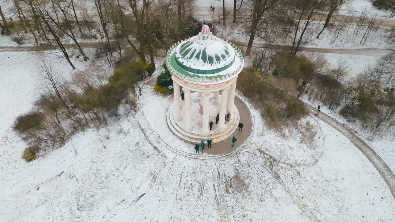 Panoramica fluida del drone che evidenzia il tempio Monopteros nel Giardino Inglese di Monaco, circondato dalla neve e che risplende nella morbida luce del sole di una mattina d'inverno.