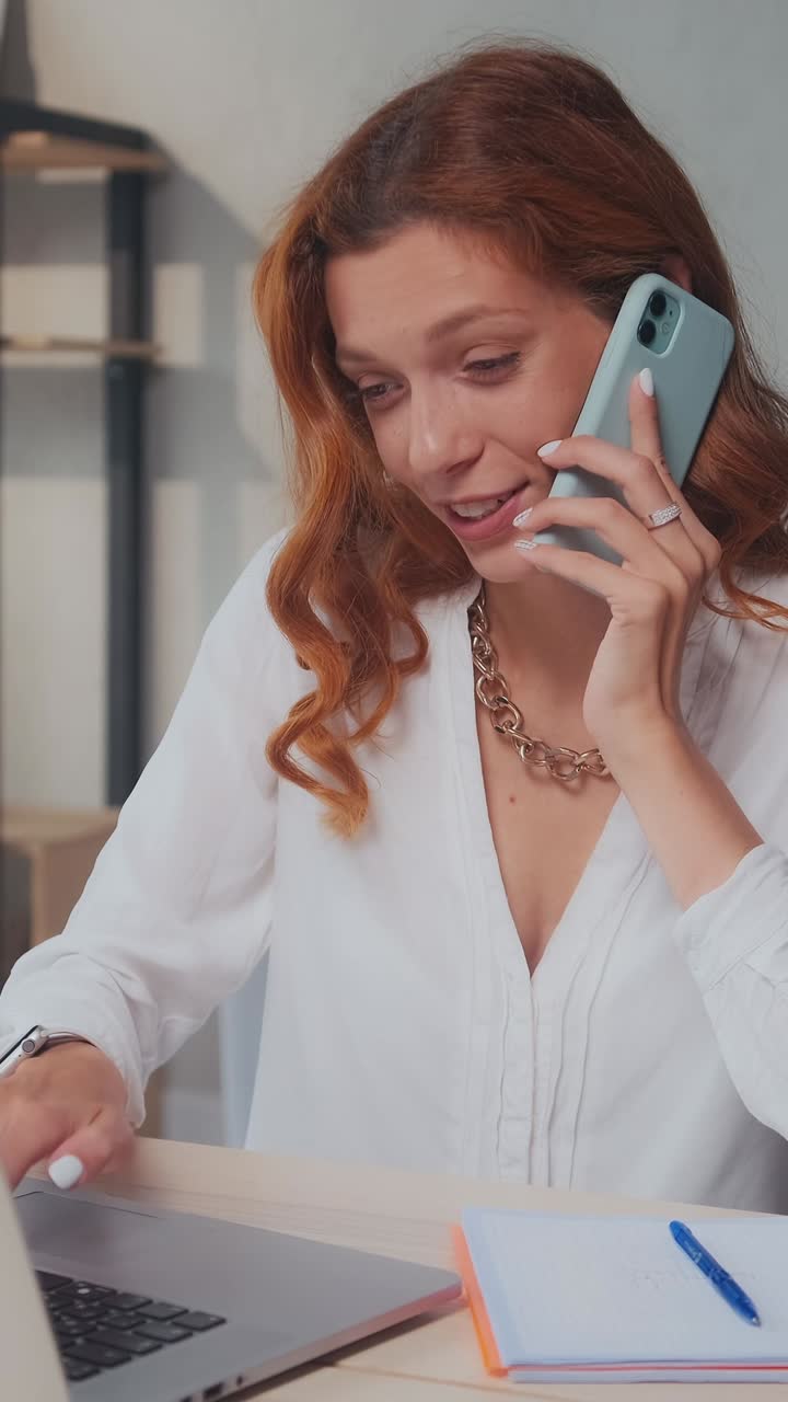 Young positive sociable caucasian woman typing on laptop and talking on phone
