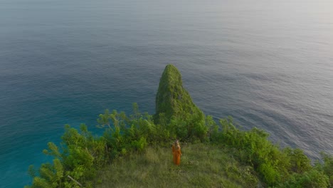 Woman-in-orange-dress-looking-in-awe-at-amazing-tropical-green-cliff-during-sunset,-Sekartaji-cliff