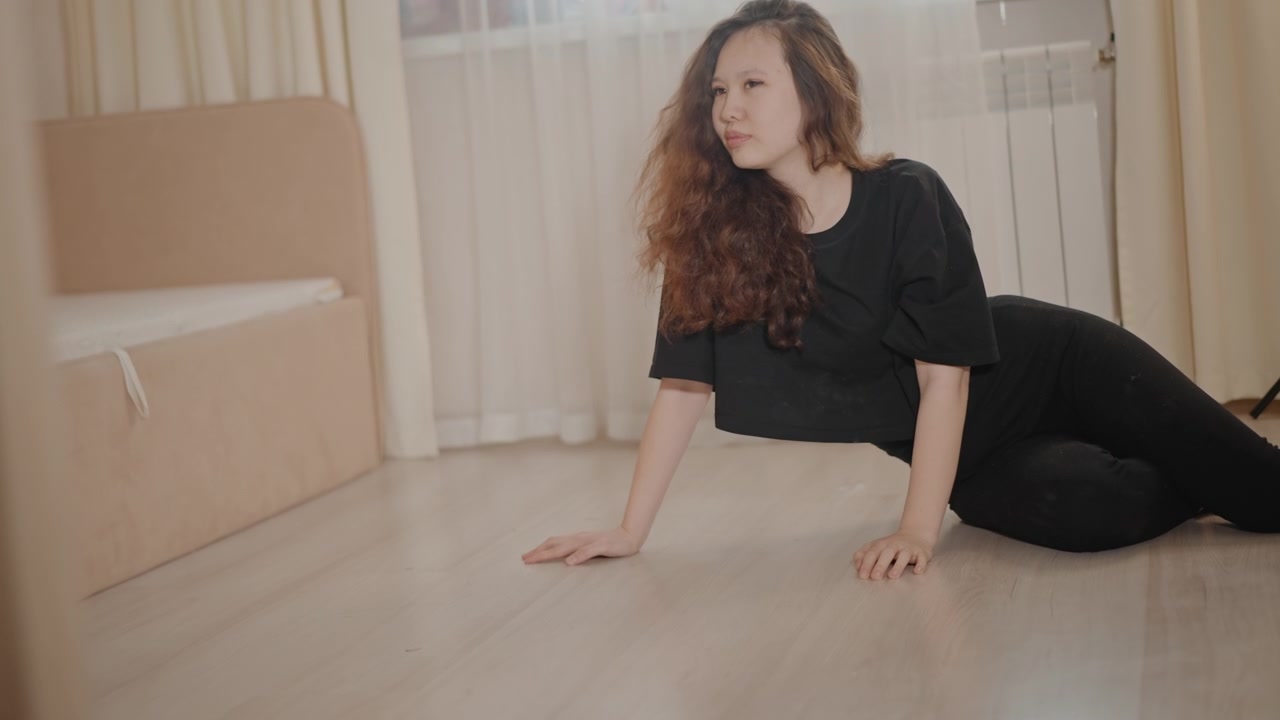 Mujer joven vestida de negro se levanta del suelo de madera clara en una luminosa habitación doméstica, elevando el torso con esfuerzo constante y girando la cabeza a la izquierda mientras pasa de una posición acostada a una sentada.