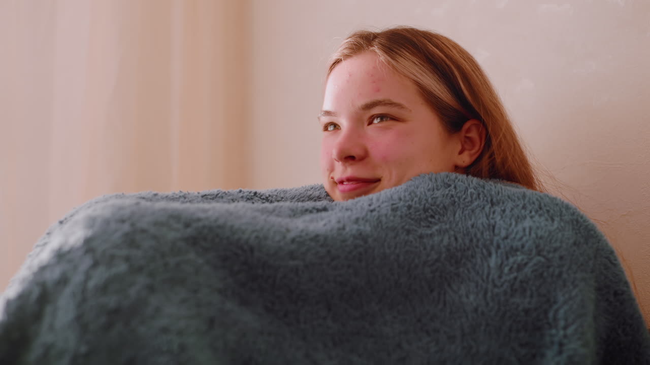 Happy college girl seated on sofa wrapping herself under blanket enjoying holiday moment with relaxed smile as sunlight enters through curtain creating cozy atmosphere of warmth and peace