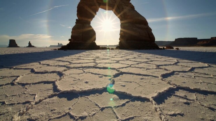 Telecamera che si abbassa rivelando un arco di roccia naturale che incornicia un'esplosione di sole per mostrare una salina crepata, con bagliore dell'obiettivo.