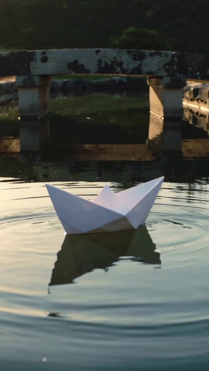 Video verticale: Barchetta di origami bianca che galleggia su uno stagno del parco spinta da una brezza, creando increspature