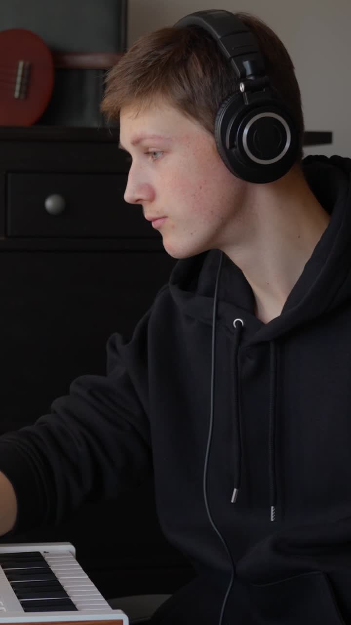 Music producer in black hoodie passionately gestures while working on MIDI keyboard in cozy bedroom studio with guitar in background