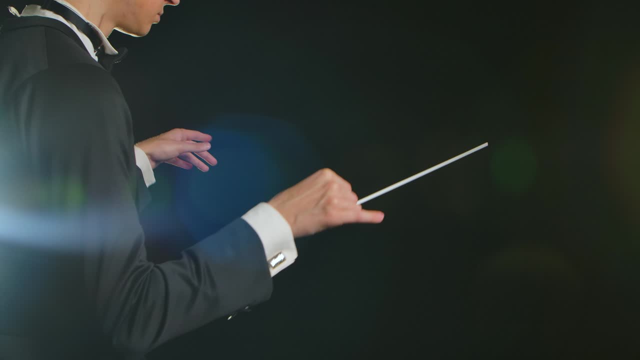 옷을 입은 교향악단 지휘자는 검은색 배경에 있는 불빛을 가진 지이의 움직임으로 음악가들을 지휘하고 있으며, 가시적인 몸짓으로 음악 공연을 지휘하고 지휘하고 있다.