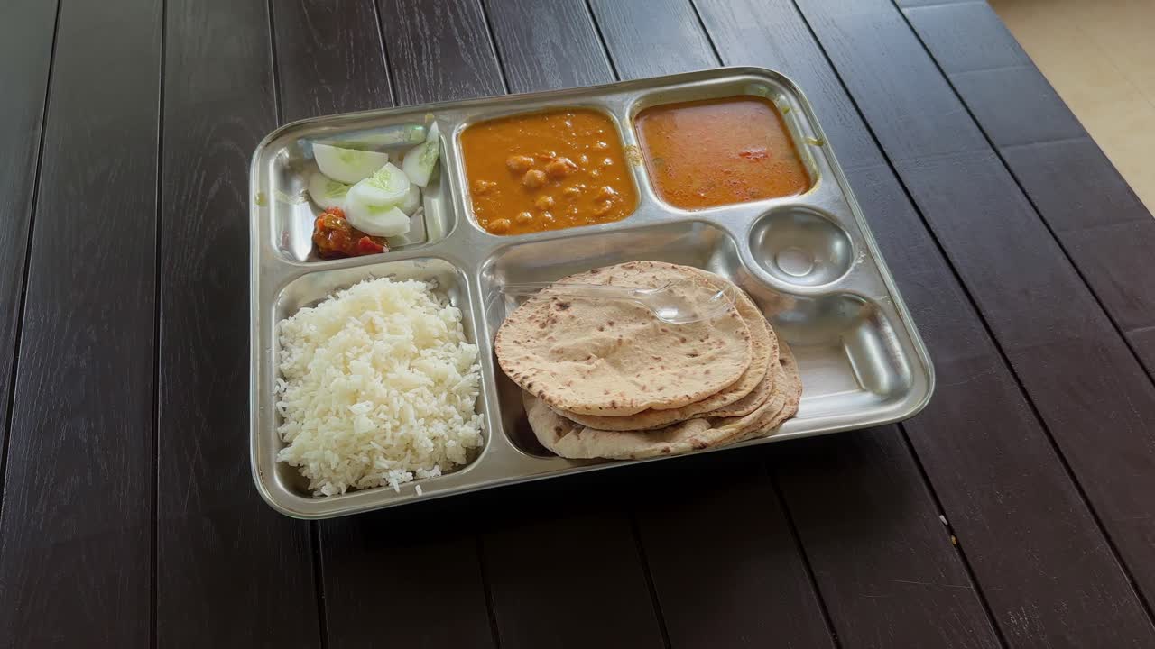 Close-up shot of a stainless steel thali containing roti, rice, dal, chickpea curry, pickle, and cucumber salad served on a wooden table