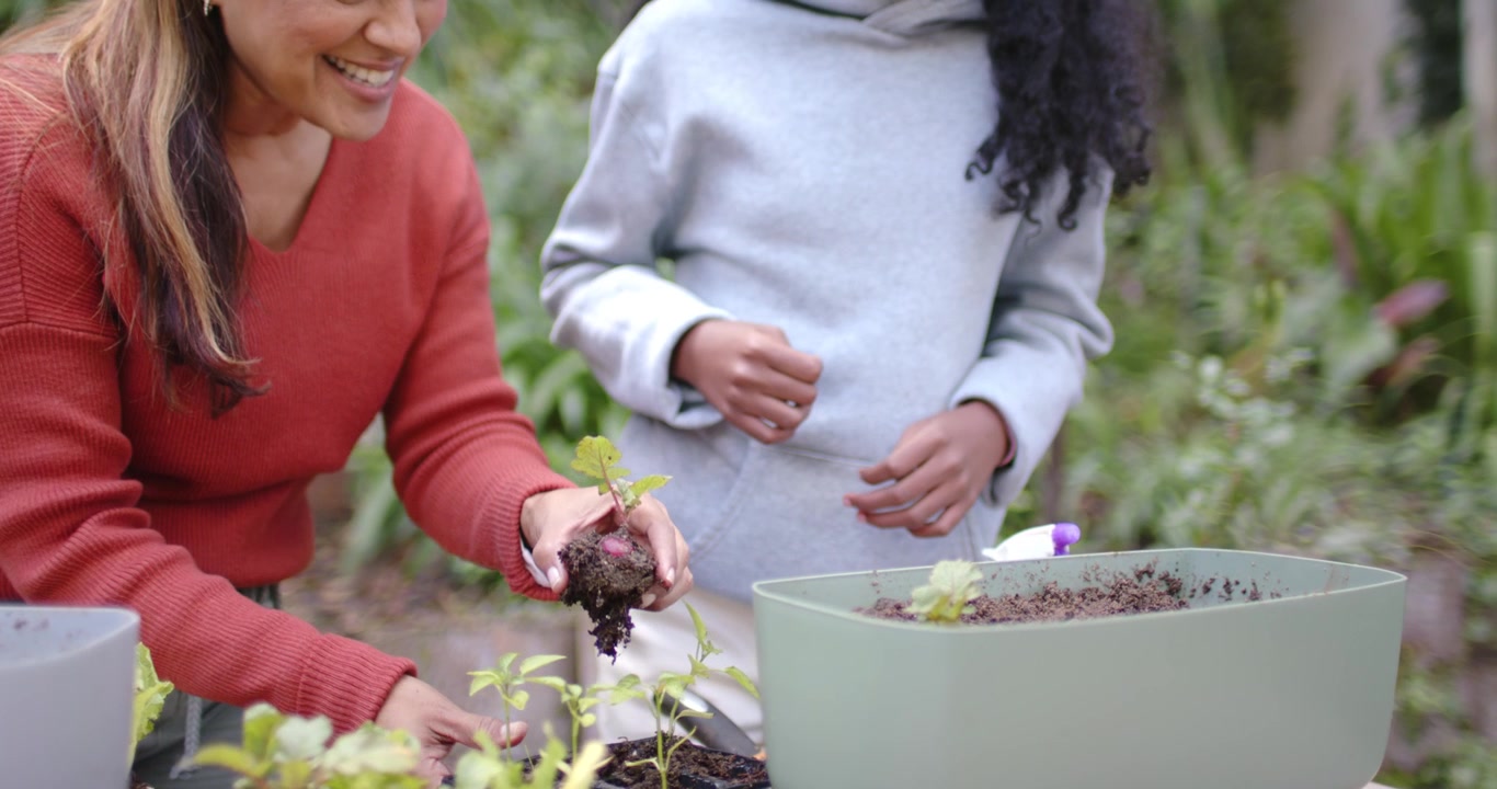 African American woman guiding girl, reaching seedling tray, planting into planter at garden table
