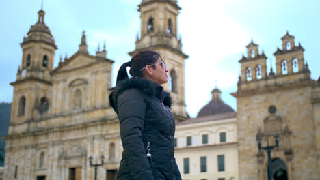 Plano-Medio-De-Una-Mujer-Vestida-Con-Una-Chaqueta-Negra-Y-Gafas-De-Sol-Azules-Mirando-El-Palacio-De-Gobierno-En-Bogotá-En-Una-Tarde-Soleada