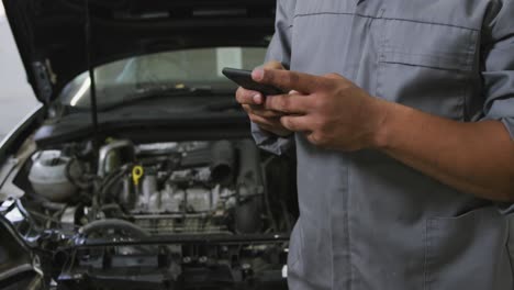 mecánico de automóviles afroamericano mirando un motor de automóvil abierto y hablando en un teléfono inteligente