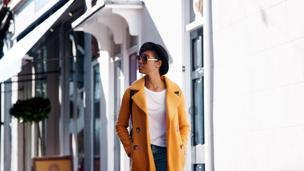Fashionable young black woman wearing blue jeans and an unbuttoned yellow pea coat walking in the street past shops on a sunny day, smiling, close up