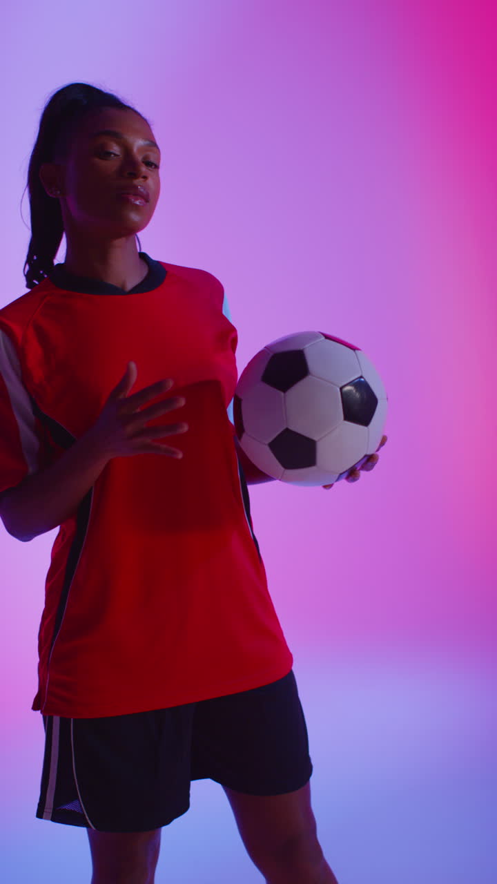 retrato vertical de estudio de video de una jugadora de fútbol o fútbol femenina con la camiseta del equipo sosteniendo la pelota bajo el brazo disparado en clave baja contra la iluminación mixta colorida