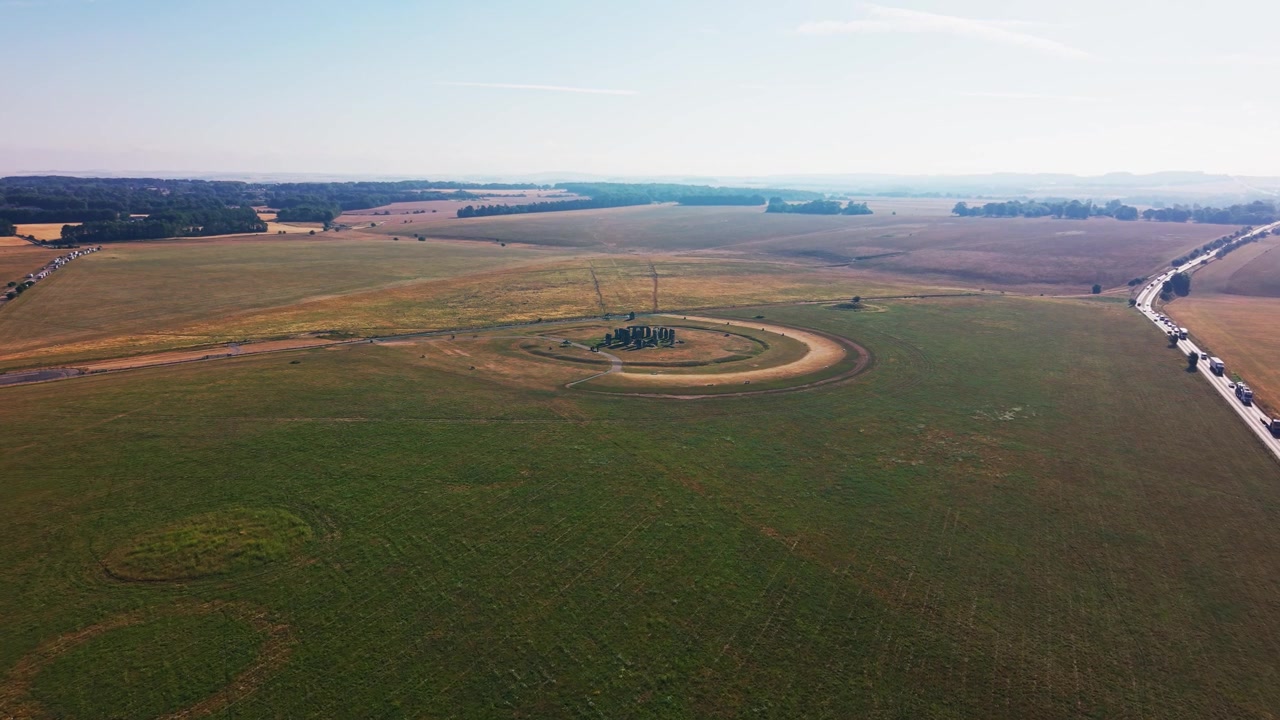 Ampie riprese aeree con drone di Stonehenge circondato da campi aperti sulla Piana di Salisbury nel Wiltshire, Inghilterra. La vista dall'alto mostra il cerchio di pietre preistorico, il flusso di visitatori e una lunga coda.