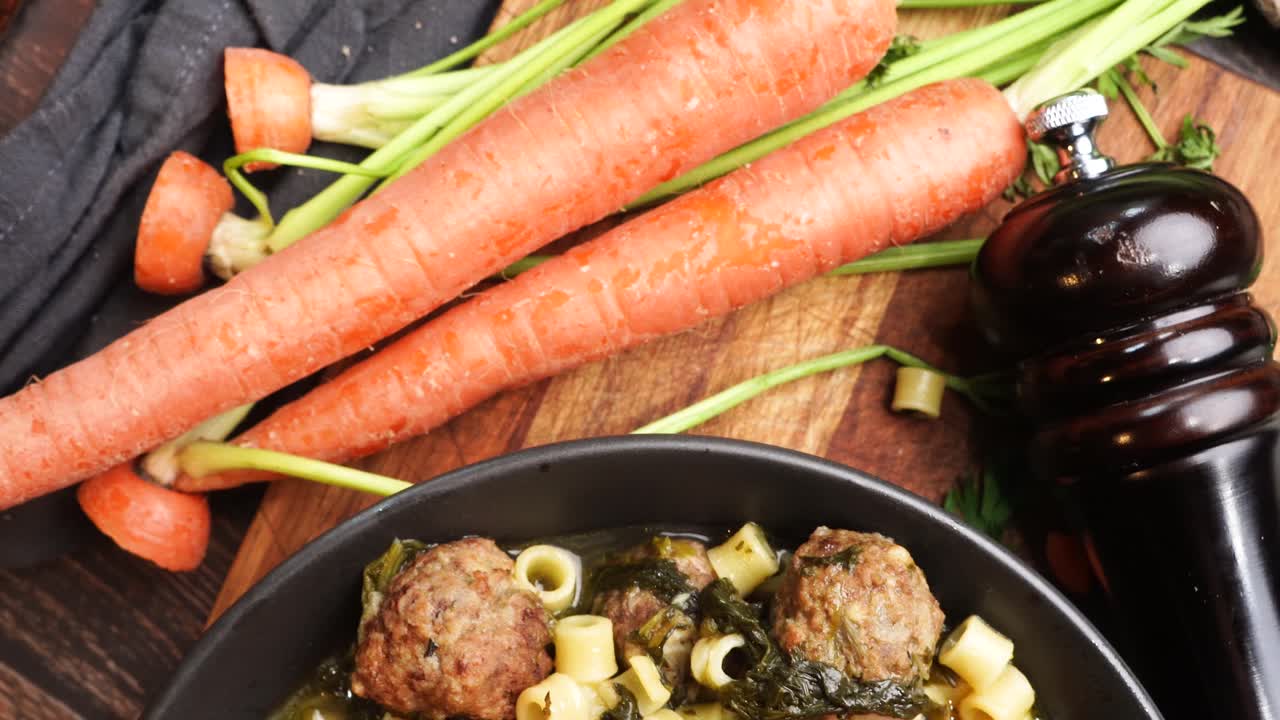 Italian Wedding Soup in a black bowl on a wooden cutting board, close up and panning across the meal. Surrounded by carrots, garlic, onions, vintage tea kettle and a pepper mill. Soup for dinner.