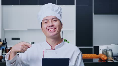 집 주방에 서 있는 동안 손에 태블릿을 가리키며 미소 짓는 아시아인 요리사