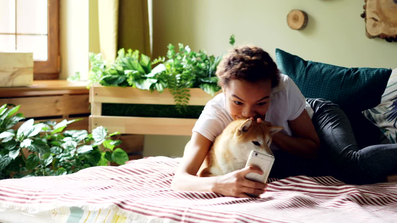 Joyful dog owner is taking selfie with cute pet lying on bed together holding smartphone posing and hugging animal. Friendship between people and puppies concept.