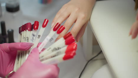 nail technician wearing pink gloves presenting various red nail polish samples to customer for selection while customer hand rests on white surface in professional nail salon during manicure session