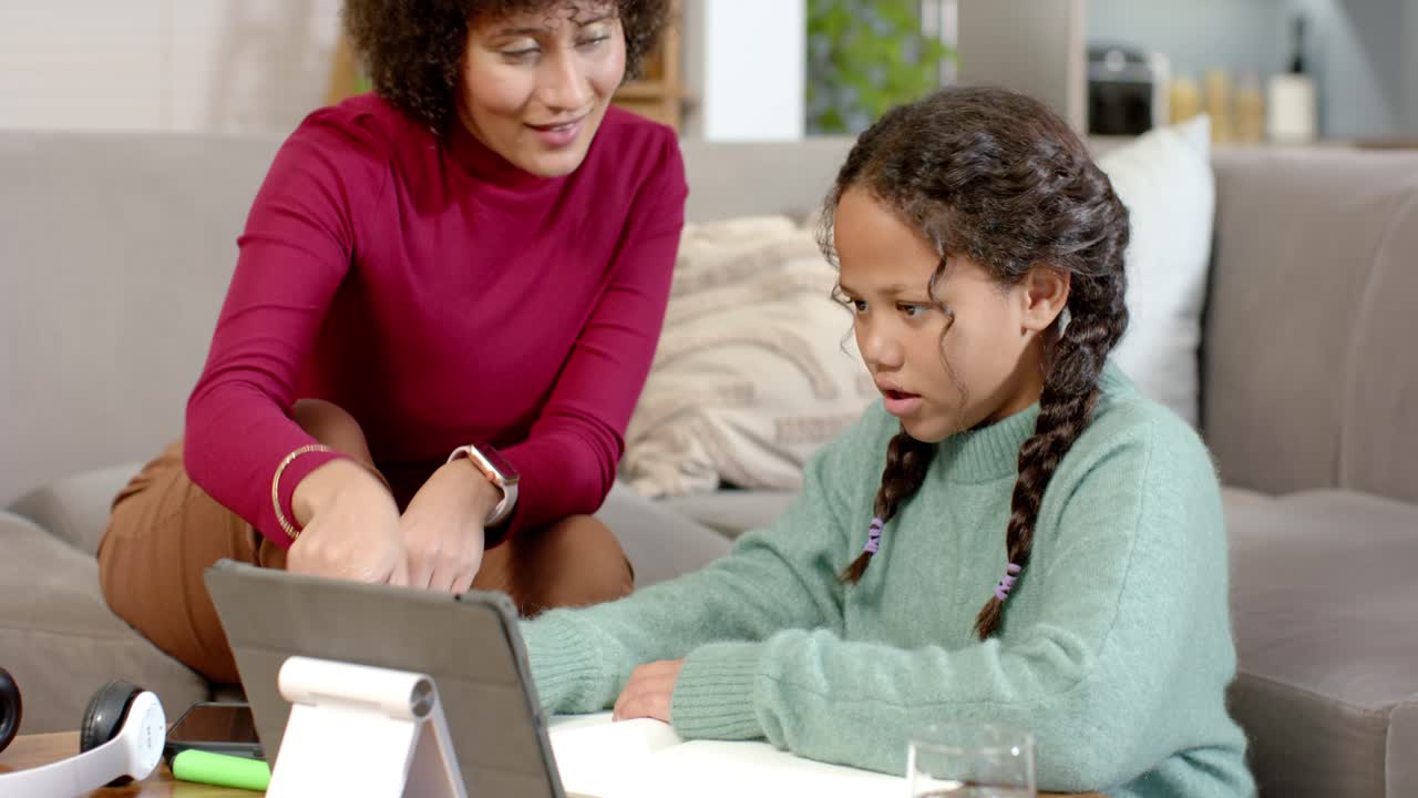 African American mom pointing at tablet at home guiding daughter tapping screen writing homework