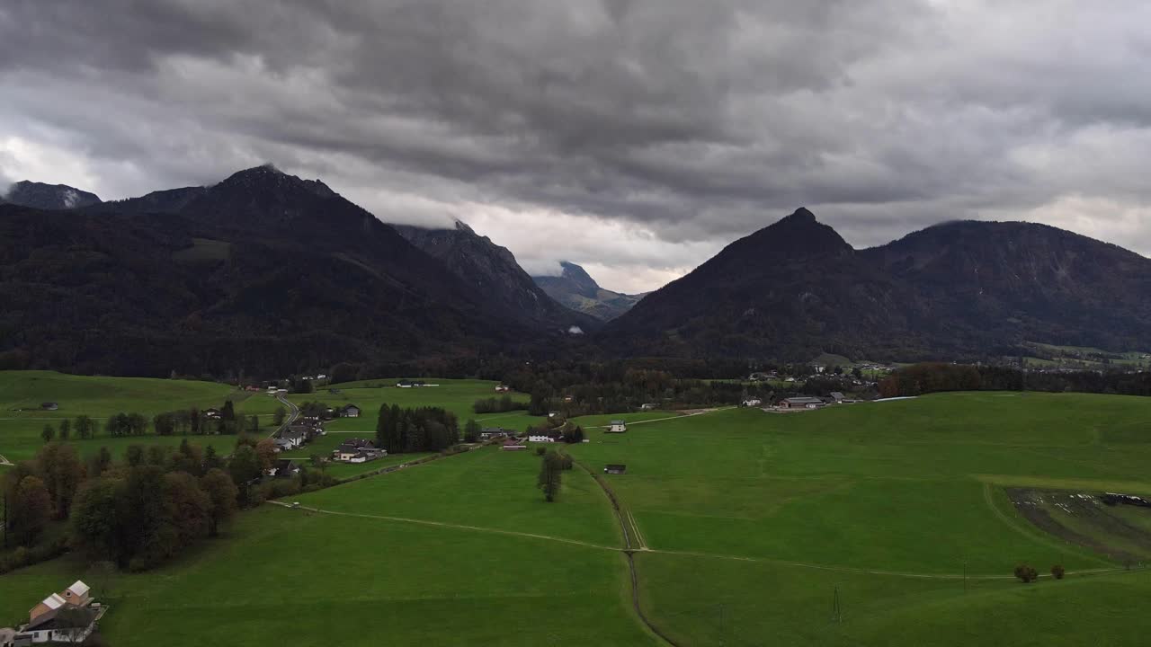 A serene rural landscape featuring a wide expanse of green fields, farmhouses, with dramatic storm clouds gathering over the distant mountain range. Flying over the green field video.